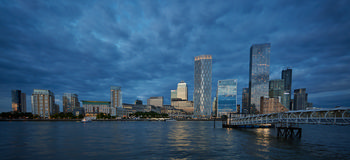 Canary Wharf Dusk Thames 2024 This urban architecture photograph captures Canary Wharf in London during the evening in the summer of 2024. The image shows the iconic skyline of Canary Wharf, with prominent modern skyscrapers such as One Canada Square rising above the city. Taken from across the River Thames, the water reflects the lights of the buildings, emphasizing the vibrant atmosphere of the area. The cloudy sky overhead adds to the overall ambiance, while the well-known London riverbank is in the foreground, highlighting the connection between the Thames and the architectural landscape of Canary Wharf at dusk.